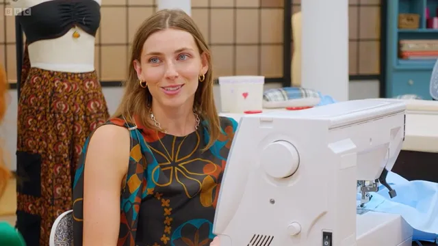Beth at her sewing machine in a sleeveless top with a bold flower print, gold hoop earrings, smiling