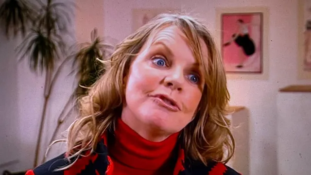 Cathy Fisher in a red polo neck and a red and black patterned jacket, in front of her paintings, mid-sentence