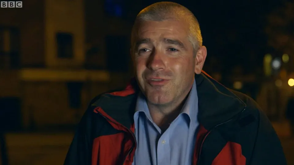 David outside at night in a red and black jacket over a blue shirt, mid-sentence