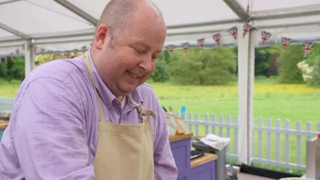 James Hillery in the Bake Off tent, smiling down at his bench, in a lilac shirt and the show's apron