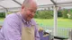 James Hillery in the Bake Off tent, smiling down at his bench, in a lilac shirt and the show's apron