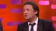 Jamie Oliver on the Graham Norton Show sofa in a dark suit and tie, in front of the orange and purple set, mid-sentence