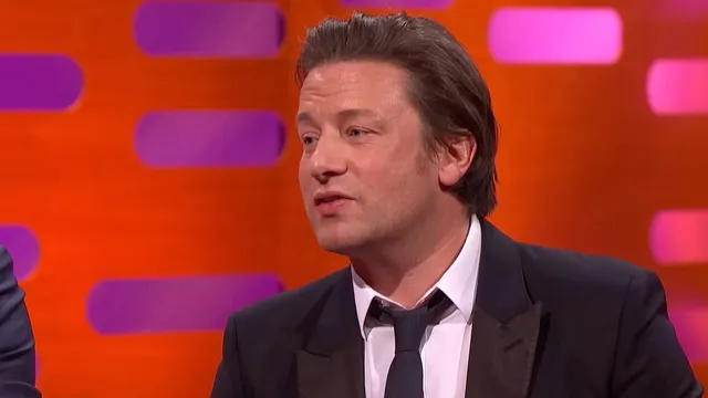 Jamie Oliver on the Graham Norton Show sofa in a dark suit and tie, in front of the orange and purple set, mid-sentence