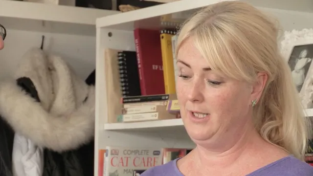 Julia in front of a bookshelf in a lilac top, a white fur hat hanging on a hook beside her, mid-sentence