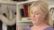 Julia in front of a bookshelf in a lilac top, a white fur hat hanging on a hook beside her, mid-sentence
