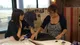 Lynda at her sewing table in a leopard-print dress and black cardigan, smiling at Claudia Winkleman, who leans on the table beside her