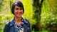 Priya O'Shea in the woods outside the tent, smiling, in a leather jacket and a zebra-print shirt