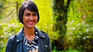 Priya O'Shea in the woods outside the tent, smiling, in a leather jacket and a zebra-print shirt