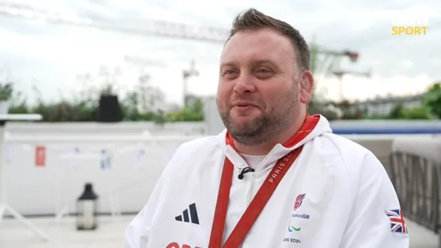 Rob Davies, smiling, in a Paralympics GB tracksuit with a red lanyard and medal, being interviewed outdoors in Paris