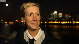 Tamara outside at night by the river, the City lights and a full moon behind her, smiling