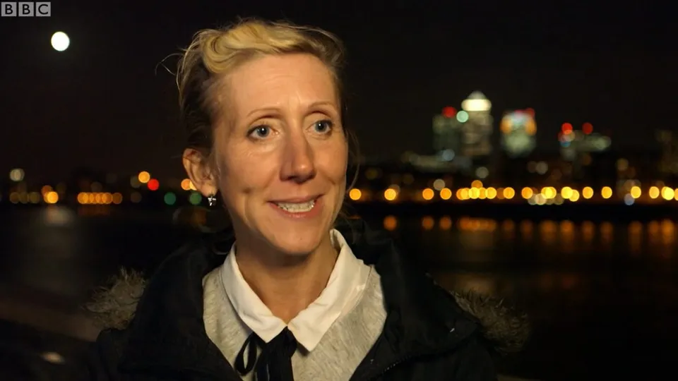 Tamara outside at night by the river, the City lights and a full moon behind her, smiling