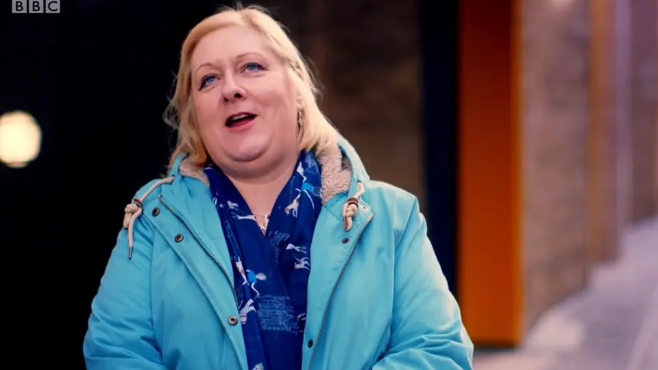 Tracey outside in a turquoise parka and a blue scarf, looking up and smiling, mid-sentence