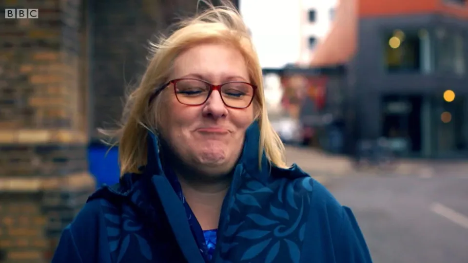 Tracey outside on a windy street in red glasses and a blue patterned coat, eyes squeezed shut and lips pressed together in a beaming smile