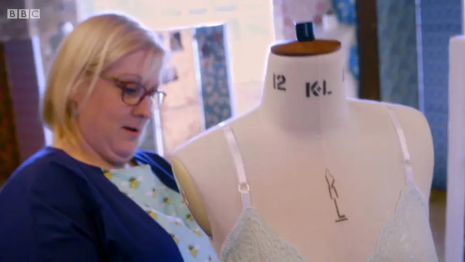 Tracey at her mannequin in glasses and a navy top over a bee-print shirt, looking down at the pale green lace bra she has made